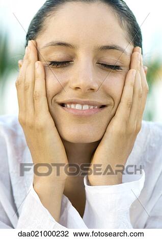 Stock Image - Woman holding face in hands, smiling, eyes closed, close-up. Fotosearch