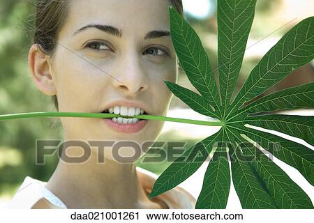 Stock Image - Woman holding large leaf in mouth, looking away. Fotosearch