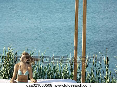 Stock Photo - Woman in bikini sitting by lake with eyes closed and hair blowing in wind. Fotosearch