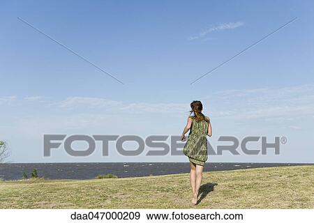 Stock Photo - Woman in dress running across grass, barefoot, rear view. Fotosearch