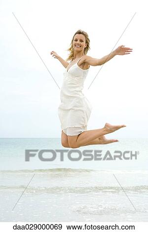 Stock Photo - Woman jumping in the air at the beach, smiling at camera. Fotosearch