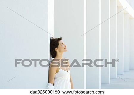 Stock Image - Woman leaning against column, eyes closed, smiling, hair tousled by breeze. Fotosearch