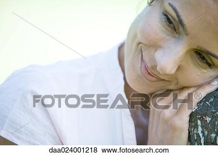 Stock Photo - Woman leaning head on wooden post, smiling, looking away, close-up. Fotosearch