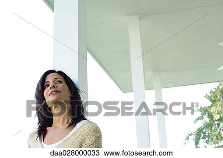 Stock Image - Woman leaning on porch column, smiling, low angle view. Fotosearch