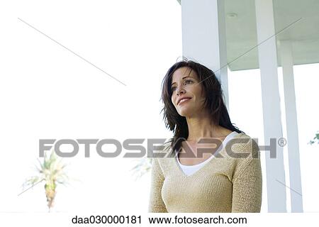 Stock Image - Woman leaning on porch column, smiling, low angle view. Fotosearch