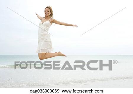 Stock Photo - Woman leaping in midair at the water's edge, arms out and legs back, smiling at camera. Fotosearch