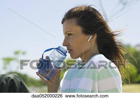 Woman listening to earphones outdoors, drinking water from bottle View Large Photo Image Stock Photograph - Woman listening to earphones outdoors, drinking water from bottle. Fotosearch