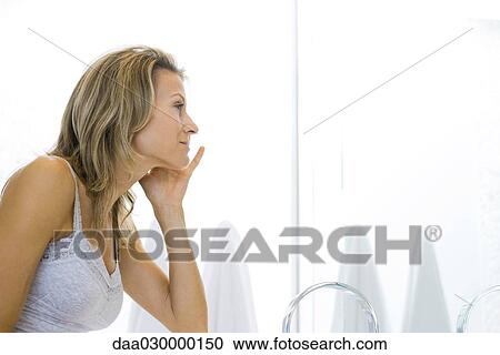 Woman looking at self in bathroom mirror, hand on face View Large Photo Image Stock Image - Woman looking at self in bathroom mirror, hand on face. Fotosearch