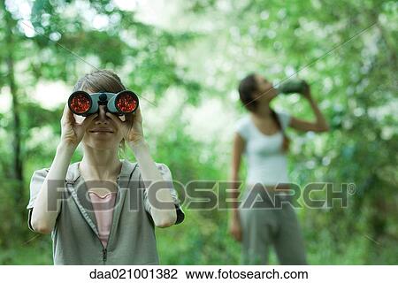 Woman looking through binoculars while friend drinks water in background View Large Photo Image Stock Image - Woman looking through binoculars while friend drinks water in background. Fotosearch