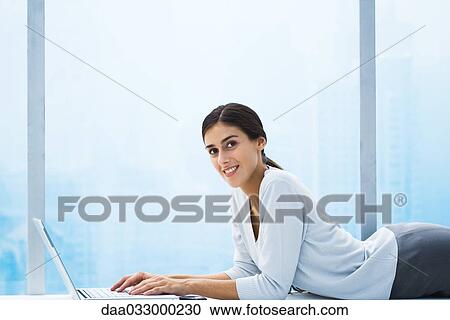 Woman lying down in front of bay window, using laptop, smiling at camera View Large Photo Image Stock Image - Woman lying down in front of bay window, using laptop, smiling at camera. Fotosearch