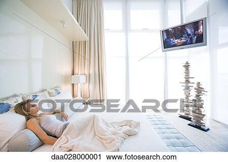 Woman lying in bed watching widescreen TV in luxurious bedroom View Large Photo Image Stock Image - Woman lying in bed watching widescreen TV in luxurious bedroom. Fotosearch