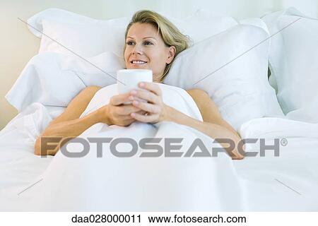 Stock Image - Woman lying in comfortable bed, holding mug, smiling. Fotosearch