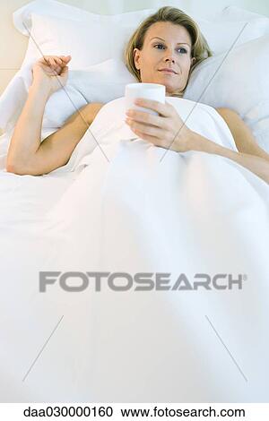 Woman lying in comfortable bed, holding mug, smiling View Large Photo Image Stock Image - Woman lying in comfortable bed, holding mug, smiling. Fotosearch