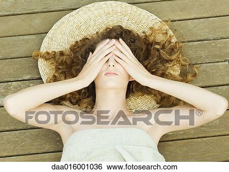 Stock Photograph - Woman lying on deck, wrapped in towel, head on mat, hands over eyes. Fotosearch