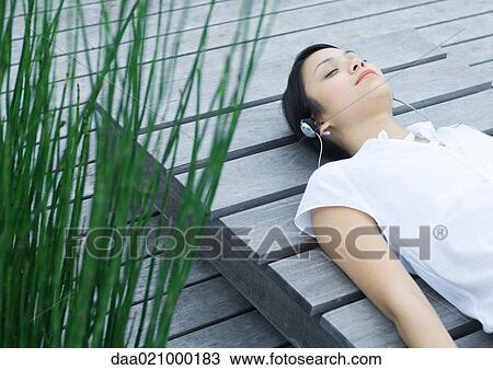 Stock Image - Woman lying on decking listening to headphones. Fotosearch