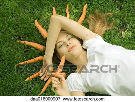 Woman lying on grass holding carrot, carrots arranged in grass around her head View Large Photo Image Stock Photo - Woman lying on grass holding carrot, carrots arranged in grass around her head. Fotosearch