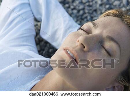 Stock Photograph - Woman lying on gravel with hands behind head, eyes closed, close-up. Fotosearch