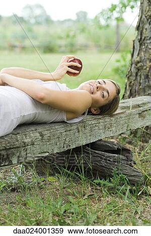 Woman lying on piece of wood, holding apple, looking at camera View Large Photo Image Stock Photo - Woman lying on piece of wood, holding apple, looking at camera. Fotosearch