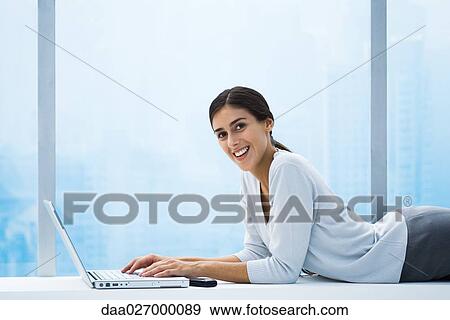 Woman lying on stomach using laptop, smiling at camera View Large Photo Image Stock Photo - Woman lying on stomach using laptop, smiling at camera. Fotosearch