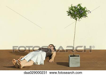 Stock Image - Woman lying on the ground beside potted tree, eyes closed. Fotosearch
