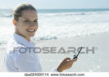 Woman on beach, holding up cell phone, looking over shoulder at camera View Large Photo Image Picture - Woman on beach, holding up cell phone, looking over shoulder at camera. Fotosearch