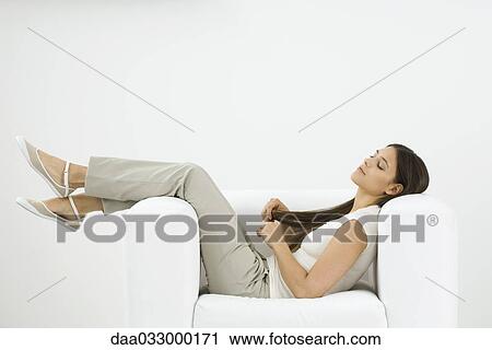 Stock Image - Woman reclining in armchair, playing with her hair. Fotosearch