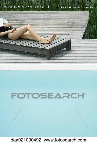 Stock Image - Woman reclining on lounge chair next to pool, sleeping with book on chest, cropped view. Fotosearch