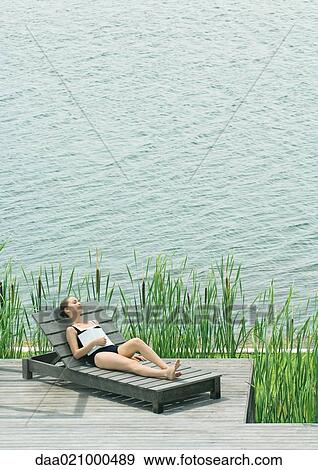 Stock Photo - Woman reclining on lounge chair next to lake, sleeping with book on chest. Fotosearch