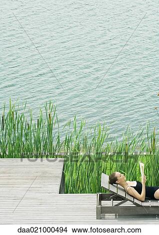 Woman reclining on lounge chair, reading, next to lake View Large Photo Image Picture - Woman reclining on lounge chair, reading, next to lake. Fotosearch