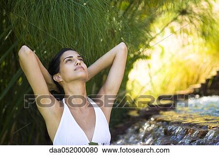 Stock Image - Woman relaxing by waterfall with hands behind head. Fotosearch