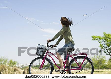 Stock Photo - Woman riding bicycle, looking at plane flying in sky. Fotosearch