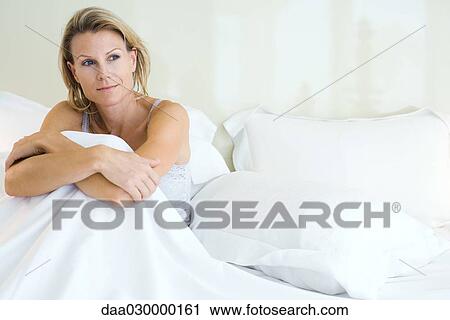 Stock Image - Woman sitting in bed, smiling, looking away. Fotosearch