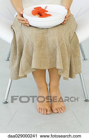 Stock Image - Woman sitting in chair, holding hibiscus in bowl of water on lap, cropped view. Fotosearch