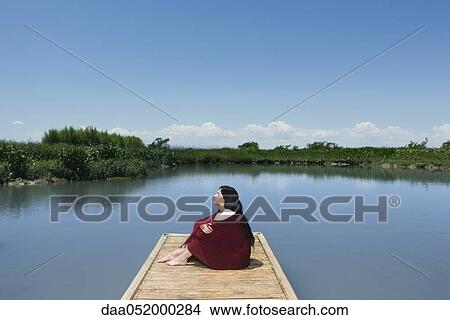 Picture - Woman sitting on dock with blanket wrapped around her, eyes closed. Fotosearch