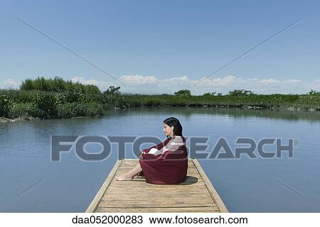 Stock Image - Woman sitting on dock with blanket wrapped around her, looking away in thought. Fotosearch