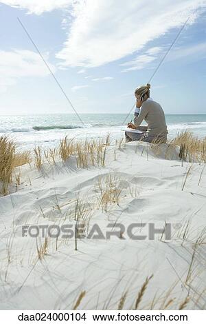 Picture - Woman sitting on dunes at beach, using cell phone and taking notes. Fotosearch