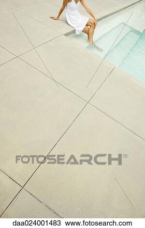 Stock Image - Woman sitting on edge of pool, feet in water, high angle view, cropped. Fotosearch