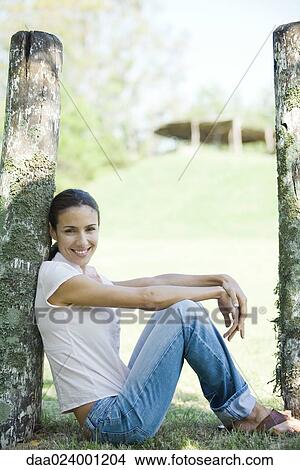 Picture - Woman sitting on grass, leaning against wooden post, full length. Fotosearch