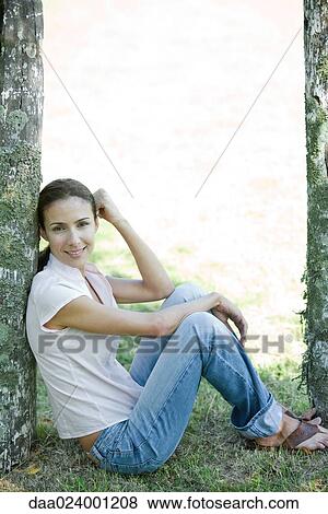 Stock Photo - Woman sitting on grass, leaning against wooden post, full length. Fotosearch