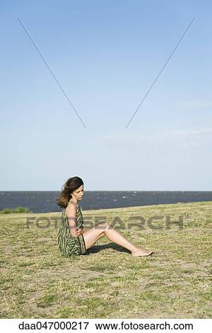 Stock Photo - Woman sitting on grass with arms folded over stomach, head down. Fotosearch