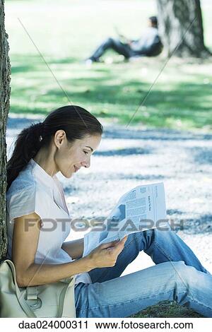 Stock Image - Woman sitting on ground in park, leaning against tree, reading newspaper. Fotosearch