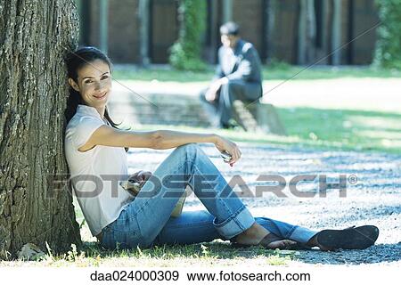 Stock Photo - Woman sitting on ground, leaning against tree, smiling at camera. Fotosearch