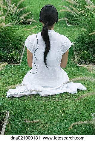Stock Image - Woman sitting on landscaped lawn, rear view, listening to headphones. Fotosearch