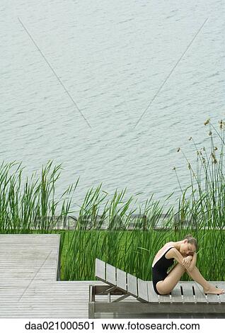 Stock Image - Woman sitting on lounge chair next to lake, hugging knees, resting head on arms. Fotosearch