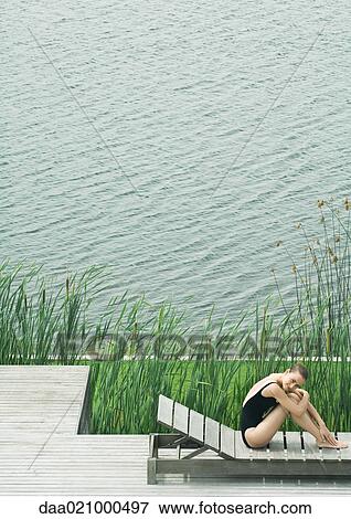Stock Photo - Woman sitting on lounge chair next to lake, hugging knees, smiling. Fotosearch