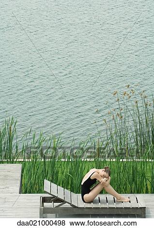 Stock Photo - Woman sitting on lounge chair next to lake, hugging knees, resting head on arms. Fotosearch