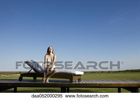 Stock Photography - Woman sitting on lounge chair outdoors, looking up dreamily. Fotosearch