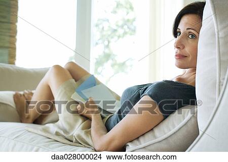 Picture - Woman sitting on sofa, holding book and stationery, looking away. Fotosearch
