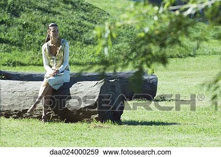 Stock Photo - Woman sitting on tree trunk, full length, looking away. Fotosearch
