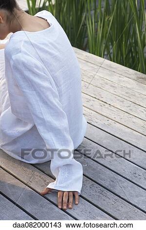 Stock Image - Woman sitting on wooden deck, cropped rear view. Fotosearch